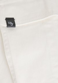 Witte denimstof met een diagonale ribbeltextuur, met een zwart leren label waarop "IBJ" is gestikt in de naad.