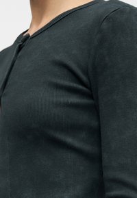 Κοντινό πλάνο ενός ατόμου που φοράει στενό μακρυμάνικο πουλόβερ με ριμπ ύφανση σε σκούρο χρώμα και μικρή λεπτομέρεια λαιμόκοψης με άνοιγμα.