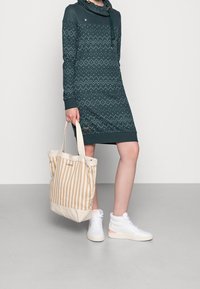 Dunkelgrünes Kleid mit Schalkragen, mit weißen geometrischen Mustern. Das Model hält eine beige gestreifte Tragetasche und trägt weiße High-Top-Sneaker.