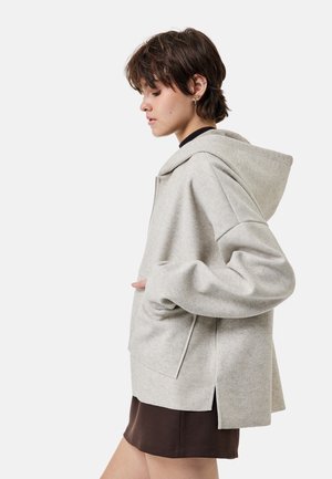 Jong persoon in lichtgrijze oversized hoodie en donkerbruine rok, naar links gericht met één hand in de zak, effen lichte achtergrond.