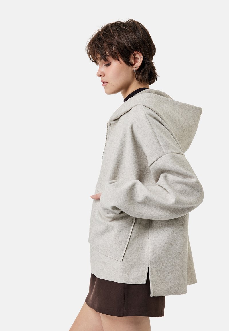 Jeune personne portant une veste à capuche oversize gris clair et une jupe marron foncé, tournée vers la gauche avec une main dans la poche, fond clair uni.