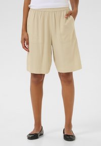 Shorts beiges avec une taille élastique, comportant deux poches latérales, une coupe ample et une texture lisse et douce. Portés avec des ballerines noires.