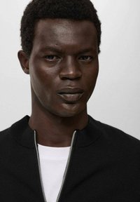 Primer plano de un hombre con pelo rizado corto que lleva un suéter negro con cremallera sobre una camisa blanca, frente a un fondo liso claro.