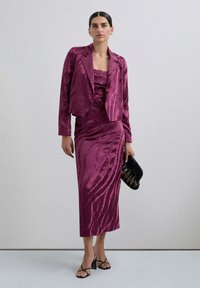 Chaqueta de traje de satén fucsia combinada con un vestido ajustado a juego que presenta patrones ondulados y un corte a la altura de la rodilla, complementado con un bolso negro.