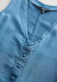 Denim gornji dio svijetloplave boje s V-izrezom, s prednjim zatvaranjem na dugmad i mekanim, teksturiranim materijalom. Jednostavan dizajn bez uzoraka.