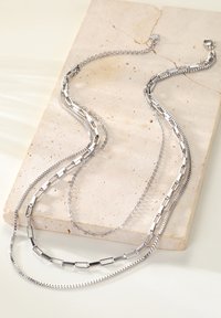 Collier argenté à plusieurs strates présentant différents styles de chaînes : designs plats, à maillons et texturés, reposant sur une surface en pierre de couleur claire.