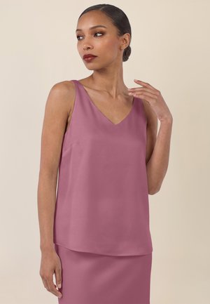 Woman with dark hair in a sleek bun wearing a sleeveless mauve top and matching skirt, looking slightly to the side with one hand near her collarbone.