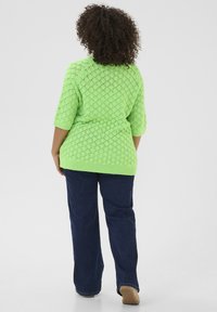 Groene gebreide trui met een ruitpatroon, korte mouwen en een losse pasvorm, gecombineerd met donkerblauwe jeans en lichtgekleurde schoenen.