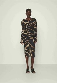 Žena v čiernych šatách s dlhým rukávom, po kolená, ktoré majú béžový tie-dye vzor a čierne sandále na vysokom opätku, stojí na jednoduchom pozadí.