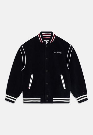 Giacca varsity in velluto blu navy con colletto a costine a strisce bianche e rosse, piping bianco, due tasche laterali e chiusura con bottoni neri.