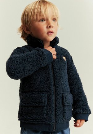 Blond kind draagt een navyblauwe fleecejas met voorzakken, kijkt iets omhoog terwijl hij met één hand de kraag van de jas vasthoudt.