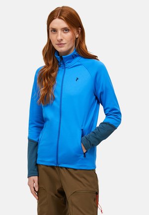 Blauwe zip-up jas van zacht materiaal met een hoge kraag. Bevat donkerblauwe accenten op de mouwen en zijzakken. Logo zichtbaar op de kraag.