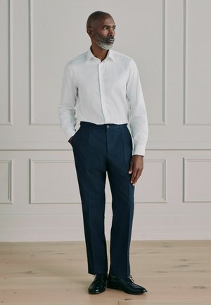 Mann mit grauem Bart, der ein weißes Hemd, marineblaue Plisseehose und schwarze Lederschuhe trägt und mit teilweise in den Taschen stehenden Händen posiert.