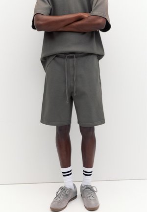 Homme debout, les bras croisés, portant un short gris, un t-shirt gris assorti, des chaussettes blanches avec des rayures noires et des baskets grises sur fond blanc.