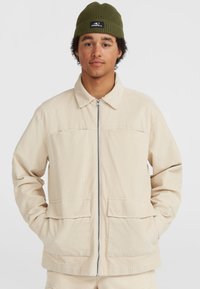 Beige corduroy jas met een kraag, een ritssluiting aan de voorkant en twee grote voorzakken. Het model draagt een olijfgroene beanie.