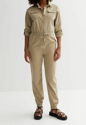 Kvinde iført beige langærmet jumpsuit med knapper og snøre i taljen samt sorte platformsandaler med ankelremme.