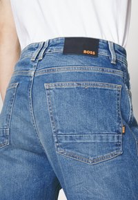 Tummansiniset farkut, joissa on korkea vyötärö. Vyötärönauhaan ja takataskuun on ommeltu oranssi "BOSS" -merkki, ja niissä on hienovaraisia ompeluyksityiskohtia.