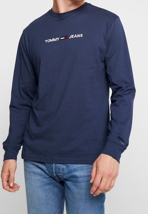 Marineblaues Langarm-T-Shirt aus Baumwollmaterial mit "TOMMY JEANS" Logo in Weiß und Rot auf der Brust. Einfacher Rundhalsausschnitt und lockere Passform.