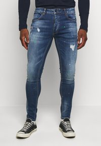 Jeans de denim azul de corte slim con detalles de desgastado y manchas de pintura. Cuenta con una cremallera, cinco bolsillos y pasadores para cinturón.
