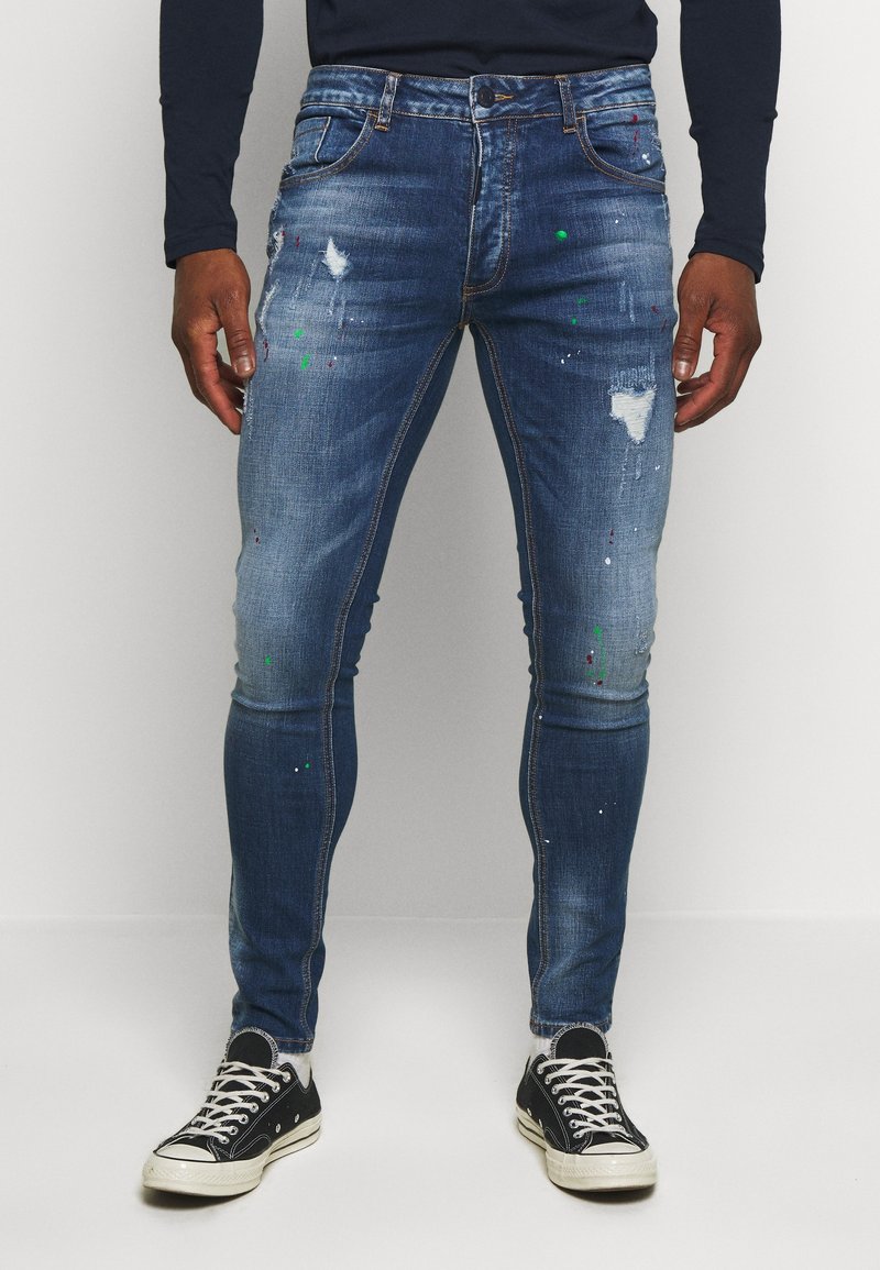 Jeans de denim azul de corte slim con detalles de desgastado y manchas de pintura. Cuenta con una cremallera, cinco bolsillos y pasadores para cinturón.