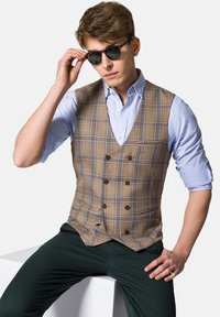 Brązowa kamizelka w kratę z podwójnym zapięciem, noszona na jasnoniebieskiej koszuli. Zielone spodnie i czarne okulary przeciwsłoneczne dopełniają wygląd.
