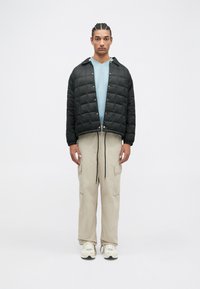 Sort quiltet jakke med trykknapper, lys blå t-shirt underneath, beige cargo bukser og hvide sneakers med sorte accenter.