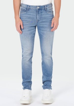 Jeans en denim bleu clair portés par une personne debout, mains détendues sur les côtés, avec des baskets blanches sur un fond blanc.