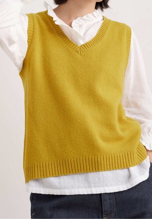 Personne portant un gilet sans manches tricoté jaune moutarde sur un chemisier blanc à volants et un jean bleu foncé.