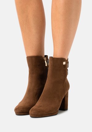 Stivaletti alla caviglia in camoscio marrone con punta quadrata, tacco a blocco e cerniera laterale. Presentano un dettaglio testurizzato sul tirante della cerniera.