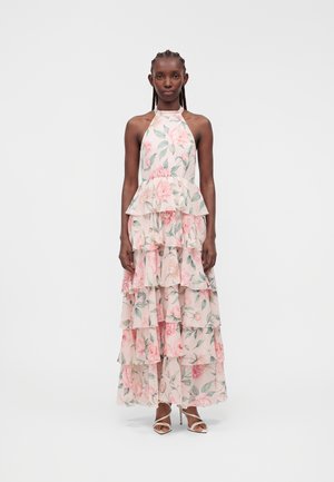 Mujer que lleva un vestido maxi sin mangas, con cuello halter y capas, con estampado floral rosa, de pie recta frente a un fondo claro y liso.
