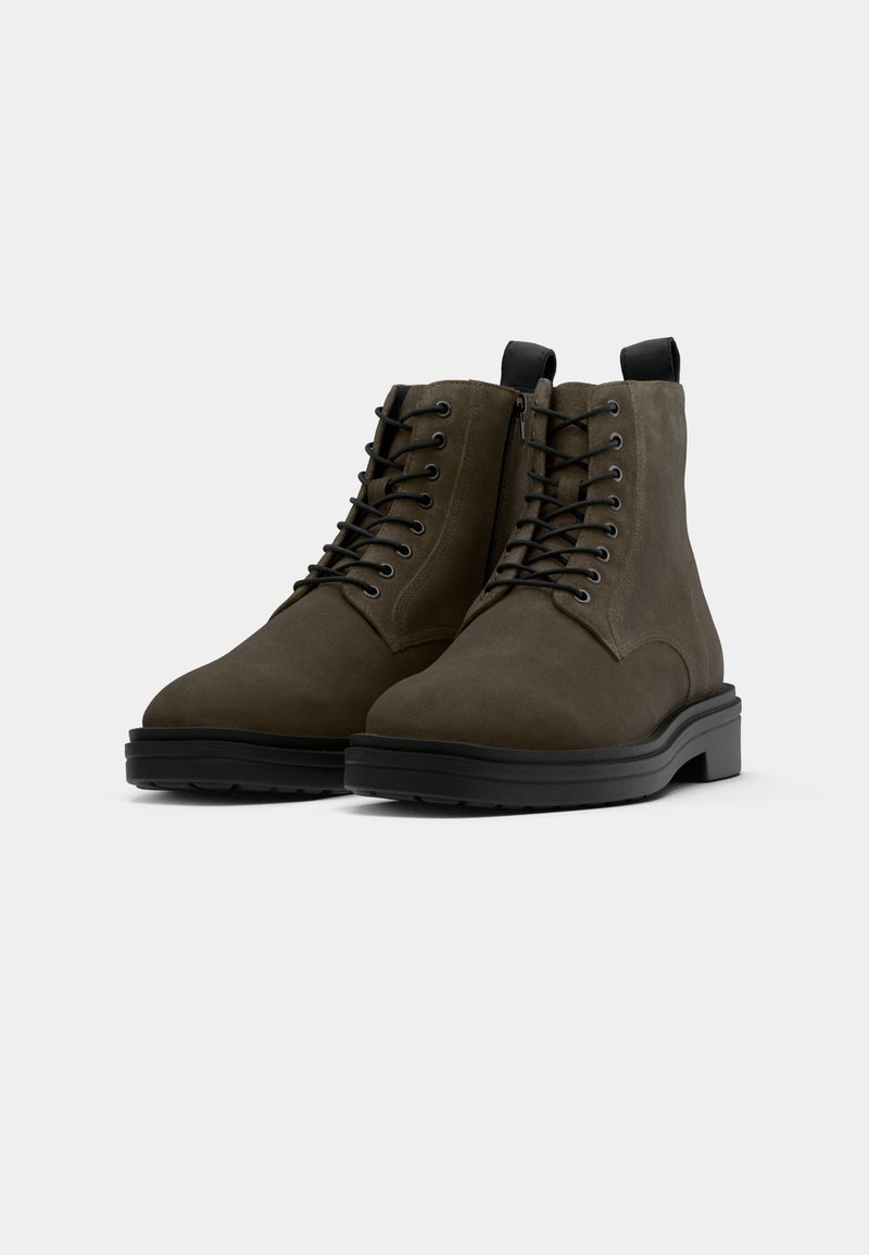 Botas de ante verde oscuro hasta el tobillo con cordones negros, punta redondeada y suela de goma negra gruesa. Cuenta con un lazo en el talón para facilitar el calzado.