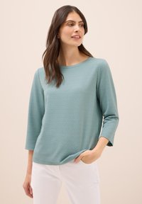 Top teal a maniche tre quarti con un motivo texturizzato, scollo rotondo e vestibilità rilassata, abbinato a pantaloni bianchi.