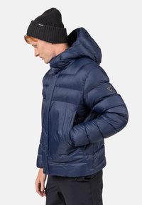 Giacca puffer blu navy con colletto alto e cappuccio oversize. Presenta sezioni trapuntate, tasche laterali e una toppa con logo sulla manica.