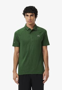 Giovane uomo con capelli ricci scuri che indossa una polo verde a maniche corte con un sottile motivo a griglia e pantaloni corti neri su uno sfondo bianco.