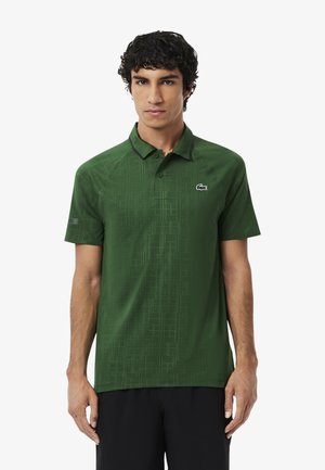 Giovane uomo con capelli ricci scuri che indossa una polo verde a maniche corte con un sottile motivo a griglia e pantaloni corti neri su uno sfondo bianco.