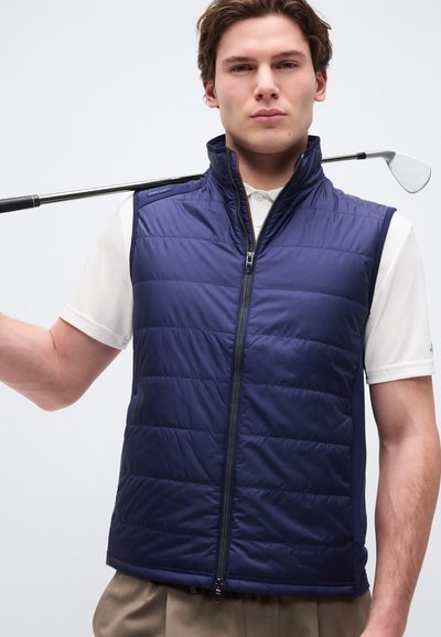 Νέος άνδρας που φοράει μπλε ναυτικό γιλέκο με ραφές και λευκό πόλο πουκάμισο, κρατά ένα γκολφ κλαμπ ακουμπώντας στους ώμους του, με φόντο έναν απλό τοίχο.