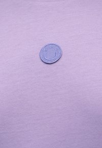 Μικρό στρογγυλό μοβ μπαλώμα με ραμμένο χαμογελαστό πρόσωπο και γράμματα σε ανοιχτό μοβ υφασμάτινο φόντο.