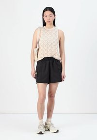 Top smanicato beige lavorato a maglia con motivo a diamante, abbinato a pantaloni corti neri con tasche laterali; il modello indossa calze bianche e sneakers.
