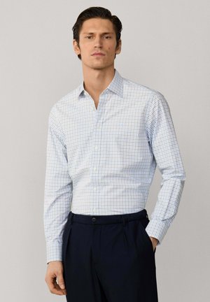 Mann trägt ein weißes Langarmhemd mit blauen und grünen Gittern, dazu dunkelblaue Hose, steht mit einer Hand in der Tasche.