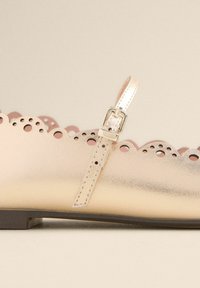 Helle goldene Leder-Schuhe mit gewellten Kanten, einem Schnallenriemen und dekorativen Ausschnitten entlang des Obermaterials. Flache Sohle.