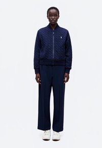 Personne debout, de face, portant une veste matelassée bleu marine zippée, un pantalon large bleu marine et des baskets blanches sur fond blanc uni.