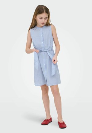 Deklica si prilagaja pas s pasom na brezrokavni modro-belo črtasti obleki, nosi rdeče ravne čevlje in stoji ob preprostem ozadju.