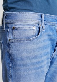 Světle modré džíny z denimu s klasickým designem pěti kapes, s kontrastním šitím, kovovými prvky a mírně vybledlou strukturou.
