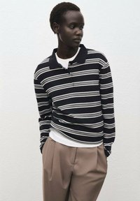 Polo a righe navy con linee bianche e grigie, indossato sopra una maglietta bianca. Abbinato a pantaloni beige larghi. Tessuto morbido.