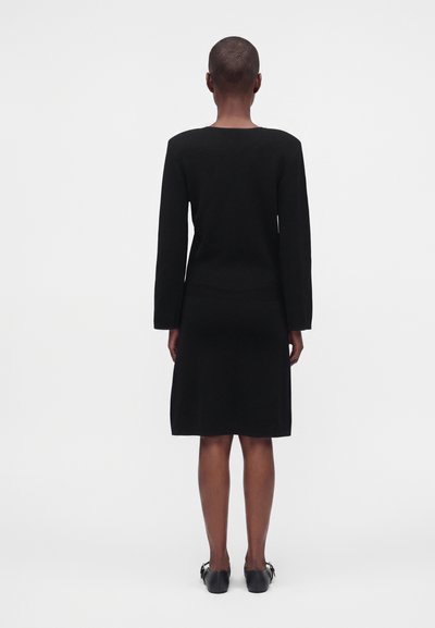 Personne aux cheveux courts portant une robe noire jusqu'aux genoux et des chaussures noires, debout, de dos, sur un fond blanc uni.