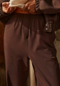 Bruine, soepele joggingbroek met een elastische tailleband en zijnaaddetails. Een bijpassend bruin bovenstuk is gedeeltelijk zichtbaar.