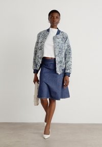 Cvetna bomber jakna v svetlo modri in beli barvi, v kombinaciji s temno modrim denim A-linijskim krilom. Model drži kremasto torbico in nosi bele pete.