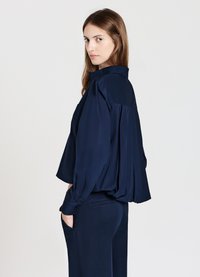 Marineblaue Satinbluse mit lockerem Schnitt, plissiertem Rücken und langen Ärmeln. Kombiniert mit passenden Hosen. Glatte Textur und minimalistisches Design.