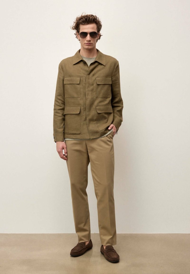 Homme portant une veste olive à col avec quatre poches à rabat, un pantalon beige, des mocassins marron et des lunettes de soleil foncées, debout contre un mur uni.