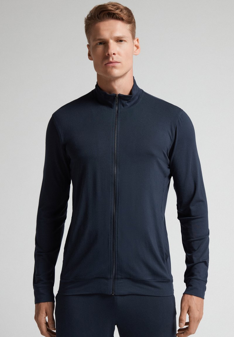 Chaqueta zip-up azul marino hecha de tela suave y ligera. Presenta un cuello alto, mangas largas y un diseño ajustado para un aspecto aerodinámico.